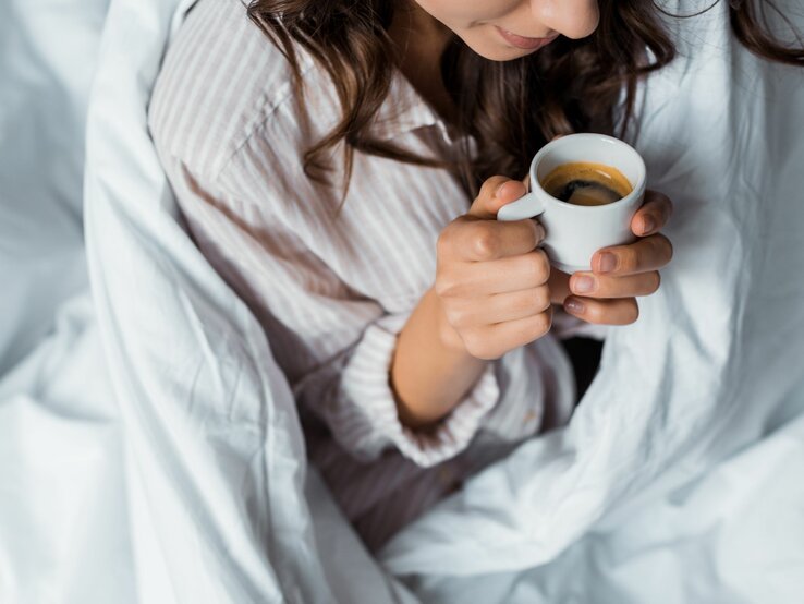 Schnappschuss einer Frau mit dunklem Haar, die morgens bequem im Bett eine Tasse Espresso trinkt.