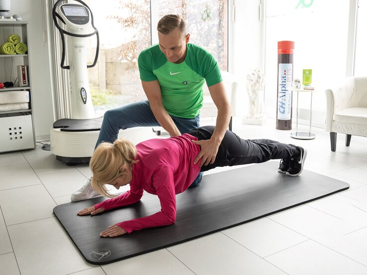 Ein Trainer in grünem Shirt hilft einer blonden Frau im pinken Pullover bei einer Plank-Übung auf einer Fitnessmatte. | © CH-Alpha Forschung