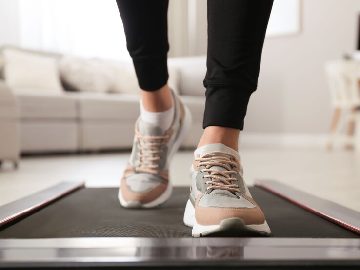 Füße in modernen Sneakers mit beige-grauen Details gehen auf einem schwarzen Walking Pad von Lidl in einem hellen Wohnzimmer