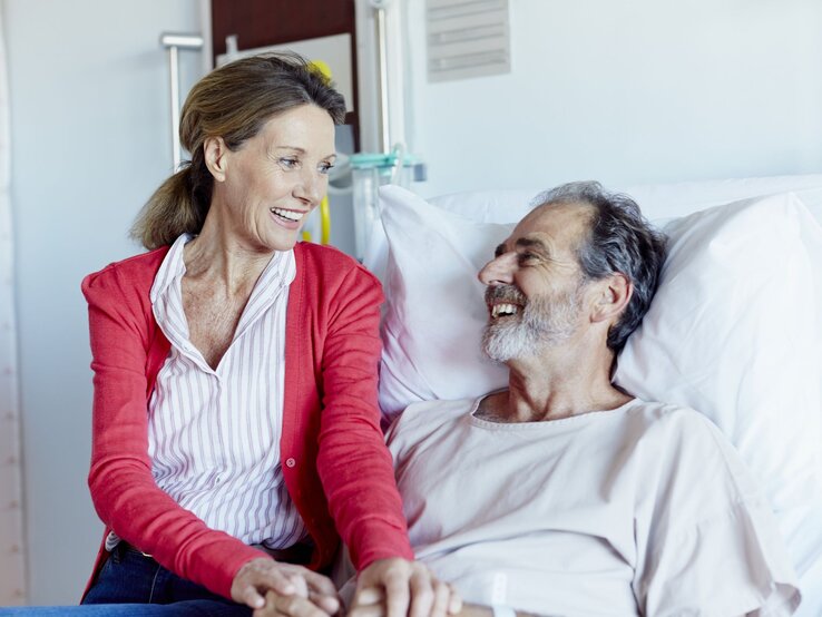 Frau, die an der Seite eines Mannes sitzt, der in einem Krankenhausbett liegt. Beide lächeln sich an und wirken entspannt und glücklich. 