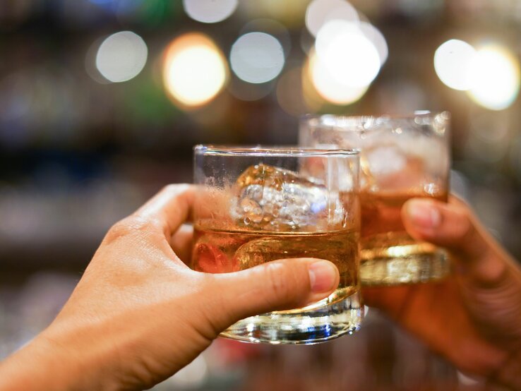 Zwei Hände stoßen mit gefüllten Whiskygläsern vor verschwommenem, buntem Hintergrund an.