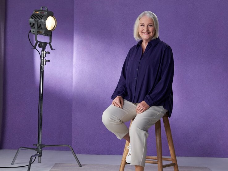 Yvonne Gallo ist eine ältere Dame, die auf einem Hocker sitzt vor einem lila Hintergrund