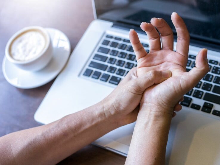 Eine Person hält sich vor einem Laptop und einer Tasse Kaffee den schmerzenden Handgelenk.
