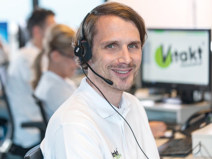 Ein lächelnder Kundenberater mit Headset in weißem Polohemd sitzt in einem modernen Callcenter, im Hintergrund verschwommene Kollegen und ein Vitakt-Logo auf dem Bildschirm. | © Vitakt