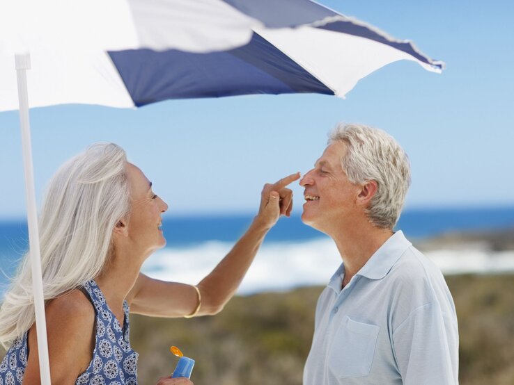 Lachende ältere Frau mit langen grauen Haaren trägt Sonnencreme auf die Nase eines ebenfalls lachenden Mannes am Strand auf.