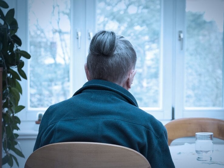 Ältere Frau, die alleine an einem Tisch sitzt, mit dem Rücken zur Kamera. Sie trägt eine blaue Jacke, und ihre grauen Haare sind zu einem Knoten hochgesteckt.