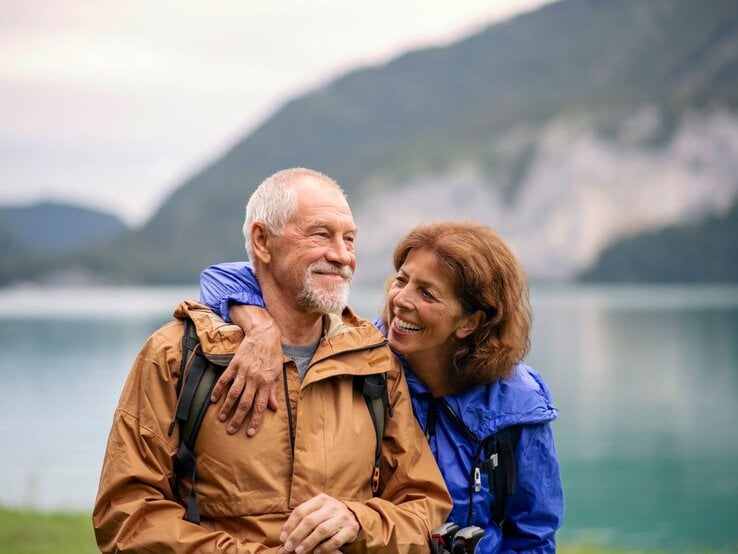 Älteres Paar in Wanderkleidung umarmt sich lächelnd vor einem Bergsee, der Mann mit grauem Bart und Frau mit braunen Haaren.