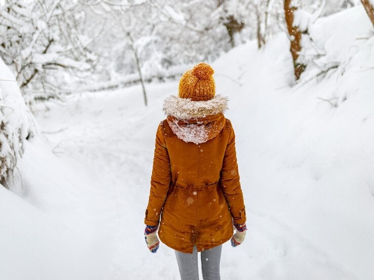 Eine Frau in warmer gelber Jacke und Mütze spaziert durch einen verschneiten Wald, während Schnee fällt. | © Adobe Stock/Shevdinov