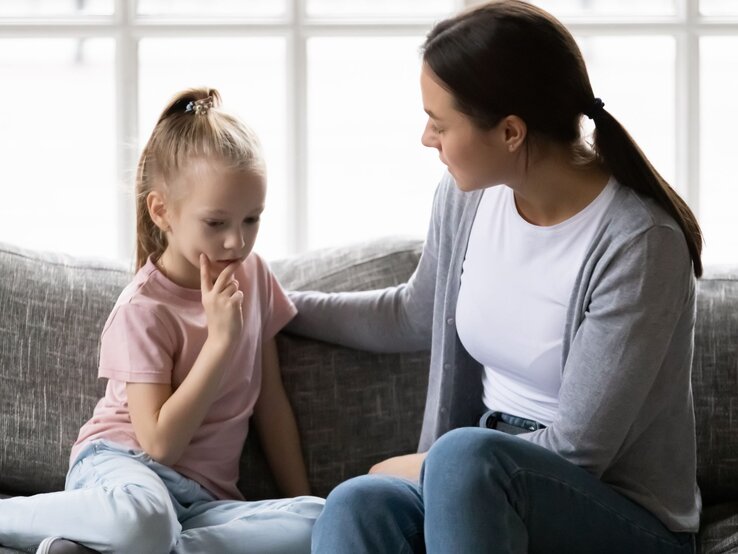 Eine junge Frau in grauem Cardigan sitzt beruhigend neben einem nachdenklichen Mädchen mit blondem Pferdeschwanz auf einem Sofa.