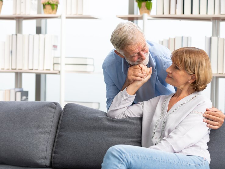 Zu sehen ist ein älteres Paar. Die Frau sitzt auf der Couch und der Mann steht hinter ihr und hält ihre Hand.