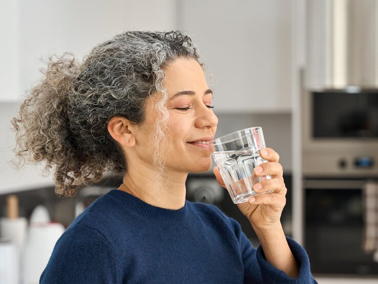 Eine Frau mit grauen Locken und blauem Pullover trinkt lächelnd ein Glas Wasser in einer modernen Küche.