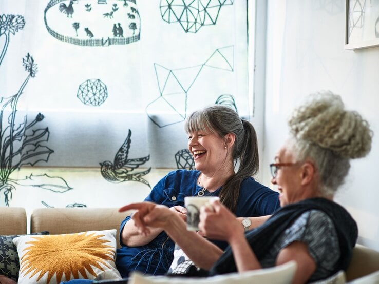 Zwei ältere Frauen sitzen lachend auf einem Sofa, eine hält eine Tasse, vor einem Fenster mit illustrierten Vorhängen. | © GettyImages/10'000 Hours