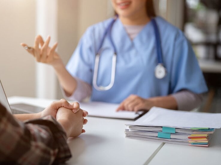 Eine junge Ärztin in blauer Arbeitskleidung spricht lächelnd mit einem Patienten, dessen Hände gefaltet auf einem weißen Tisch liegen. | © GettyImages/BongkarnThanyakij