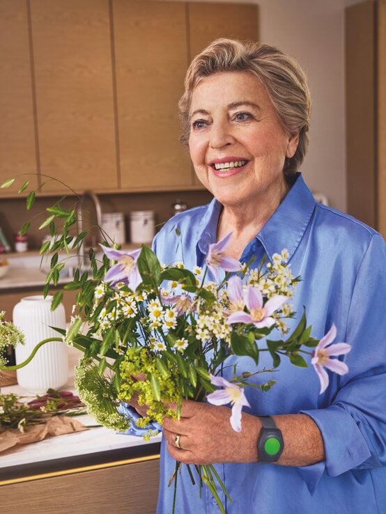 Die Schauspielerin Marie-Luise Marjan hält einen Strauß Blumen in der Hand und trägt ein Notrufarmband von Vitakt. | © Vitakt Hausnotruf