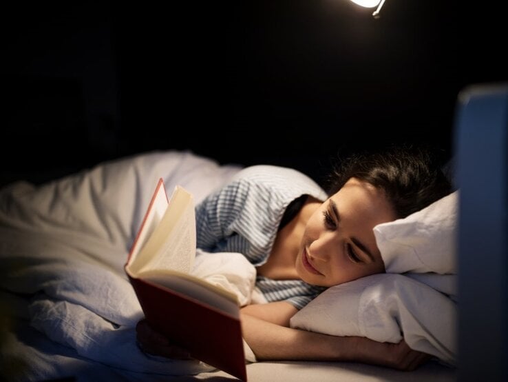 Junge Frau mit dunklen Haaren liest entspannt ein Buch im Bett, beleuchtet von einer warmen Nachttischlampe in dunkler Umgebung.