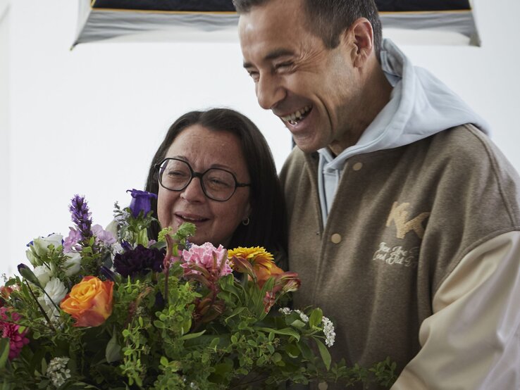Eine ältere Frau mit Brille und ein lachender Mann in beiger Jacke halten gemeinsam einen bunten Blumenstrauß.