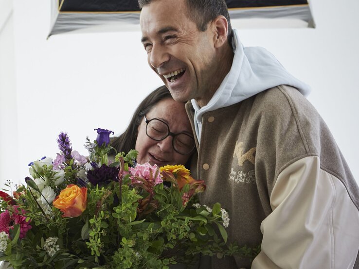 Ein lachender Mann in beiger Jacke umarmt eine ältere Frau mit Brille und dunklen Haaren, die einen bunten Blumenstrauß hält. | © dsphotos.de