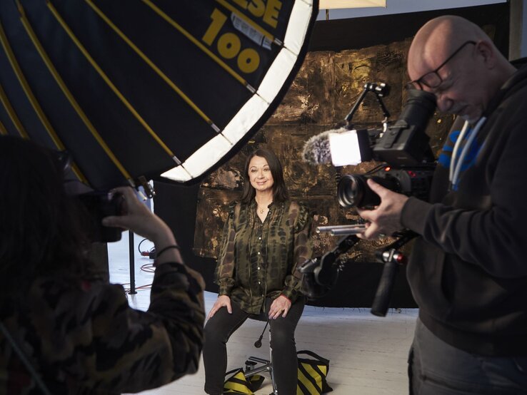 Eine Frau mittleren Alters in grünem Camouflagehemd sitzt lächelnd vor einer Kamera, während zwei Personen sie fotografieren und filmen. | © dsphotos.de