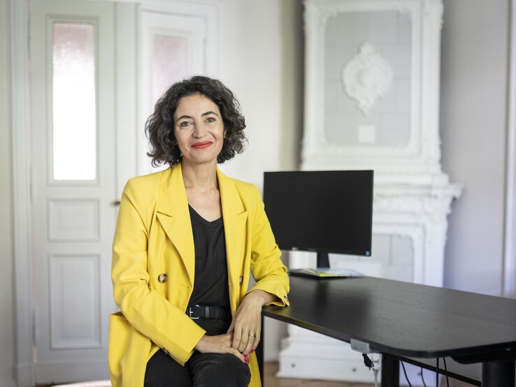 Eine Frau mittleren Alters mit lockigem dunklem Haar und rotem Lippenstift lächelt in gelbem Blazer in einem hellen Büro. | © dsphotos.de