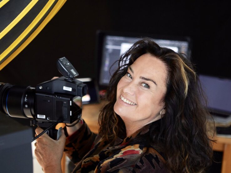 Lachende Frau mit dunklen, welligen Haaren und einem Camouflage-Mantel hält eine Kamera in einem Studio mit professioneller Beleuchtung. | © dsphotos.de