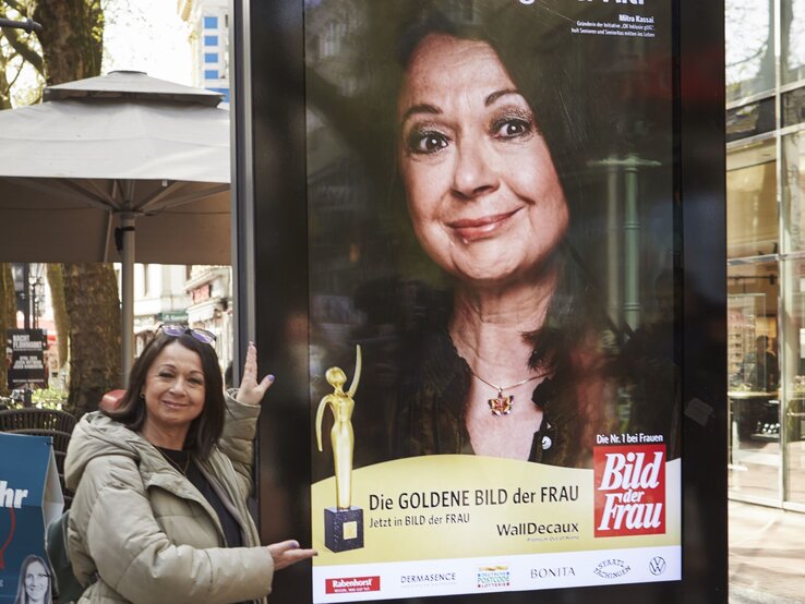 Eine ältere Frau in beiger Jacke präsentiert lächelnd ein großes Werbeplakat der "Goldene Bild der Frau" in einer belebten Fußgängerzone.