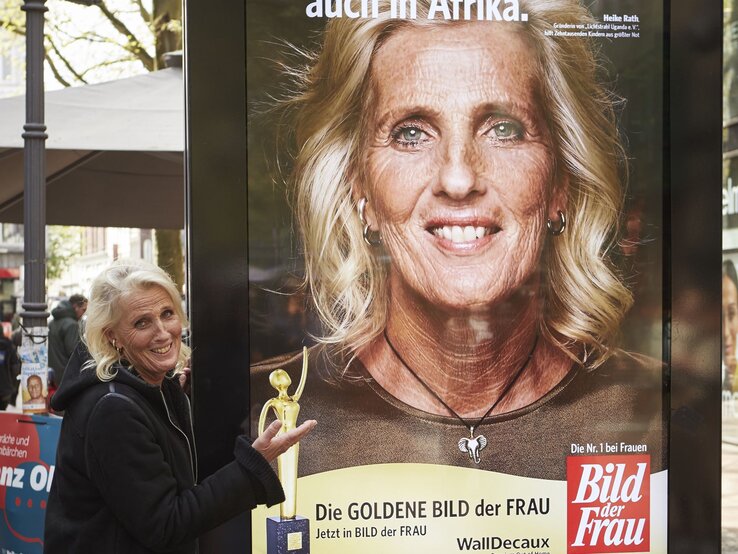 Eine ältere Frau mit schulterlangem blondem Haar und schwarzer Jacke präsentiert lächelnd ein Werbeplakat der "Goldene Bild der Frau" auf einem belebten Platz. | © dsphotos.de