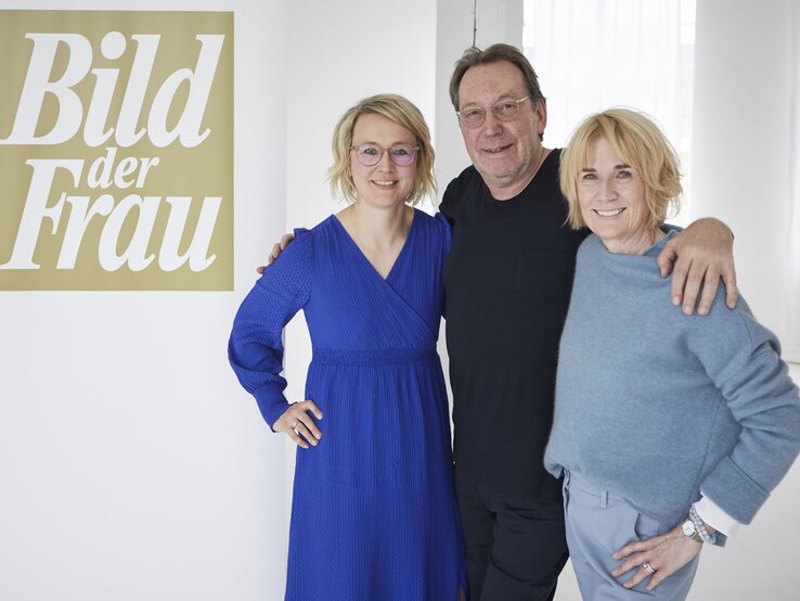 Drei lächelnde Personen stehen in einem hellen Raum vor einem "Bild der Frau"-Banner, zwei Frauen und ein Mann, Arm in Arm. | © dsphotos.de