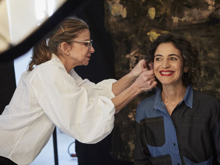 Visagistin mit Brille und weißer Bluse richtet Haare einer lächelnden Frau mit lockigem Haar und rotem Lippenstift in einem Studio. | © dsphotos.de