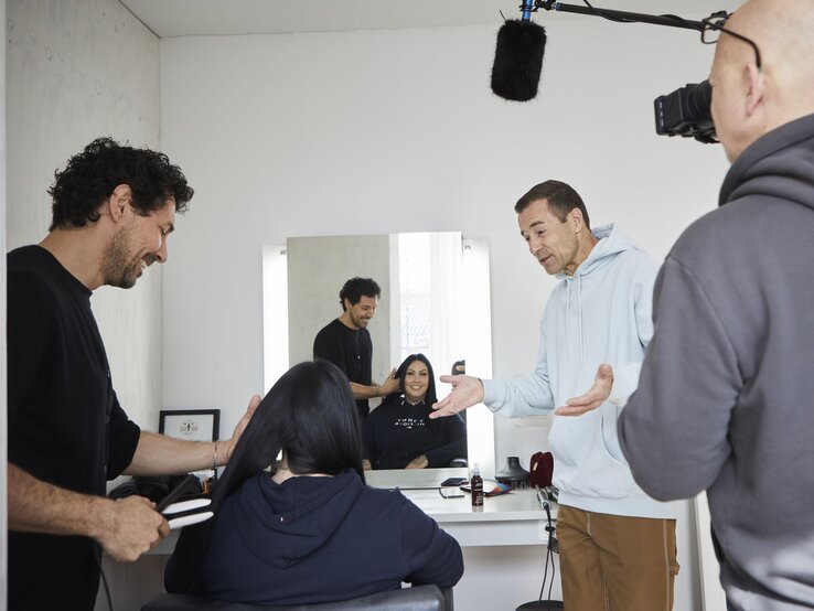 Visagist und Mann im hellen Hoodie unterhalten sich, während sie die Haare einer Frau im Friseursalon stylen und ein Kameramann aufnimmt. | © dsphotos.de