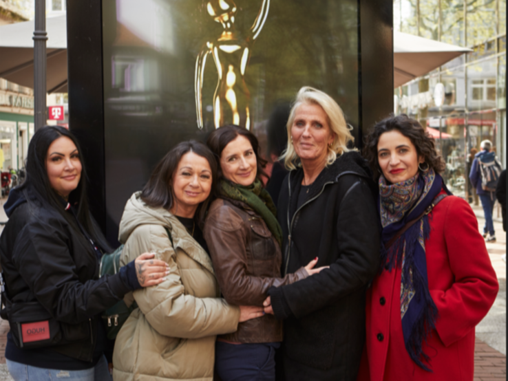 Fünf Frauen lächeln und umarmen sich in einer Fußgängerzone, vor einer Skulptur in einer Glasvitrine.