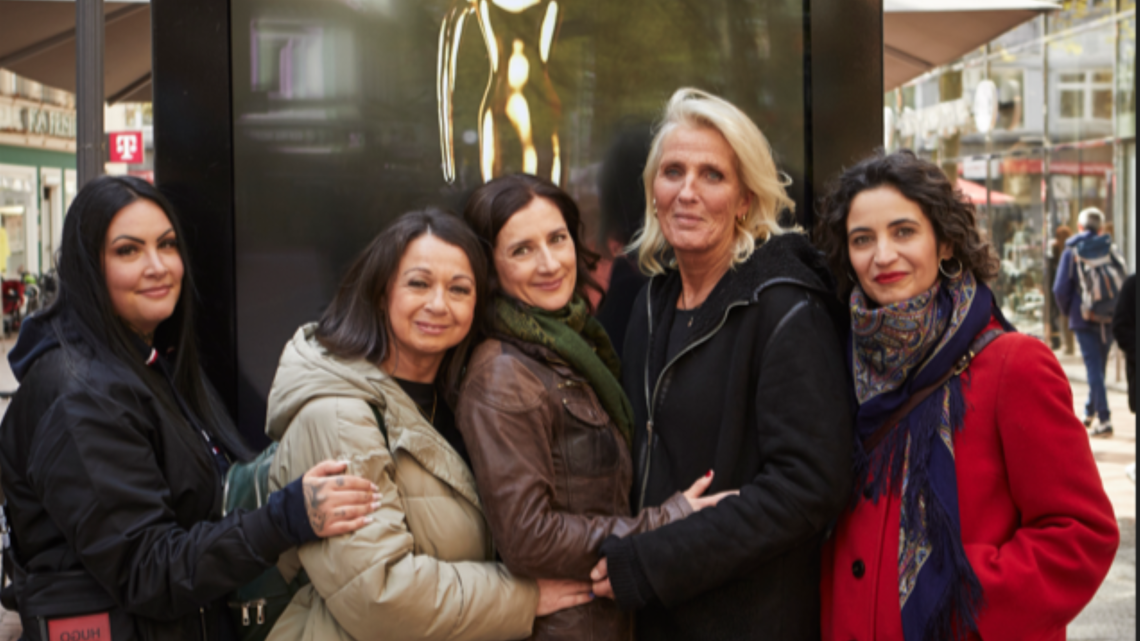 Fünf Frauen lächeln und umarmen sich in einer Fußgängerzone, vor einer Skulptur in einer Glasvitrine.