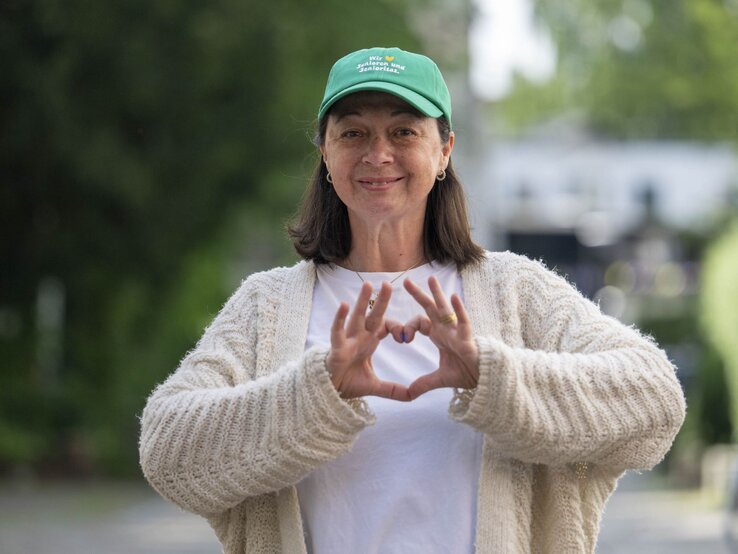 Eine lächelnde Frau mittleren Alters mit grüner Kappe und beiger Strickjacke formt ein Herz mit ihren Händen vor unscharfem Hintergrund. | © Ulrike Schacht