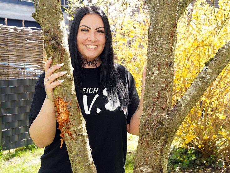 Frau mit langen schwarzen Haaren, tätowiertem Hals und schwarzem Shirt lächelt zwischen zwei Bäumen im Garten. | © Marion Vogel