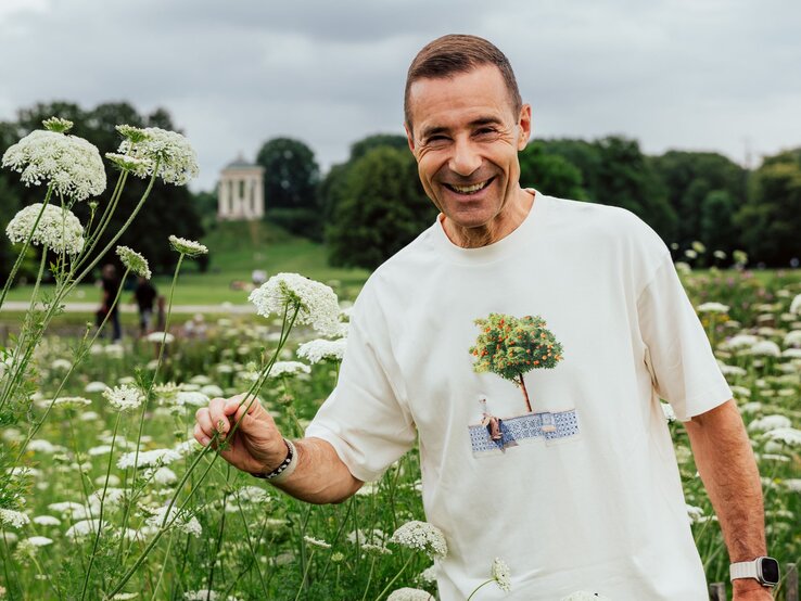 Ein lächelnder Mann mittleren Alters mit kurzen braunen Haaren steht in einer Wiese mit weißen Blüten vor einem bewölkten Himmel.