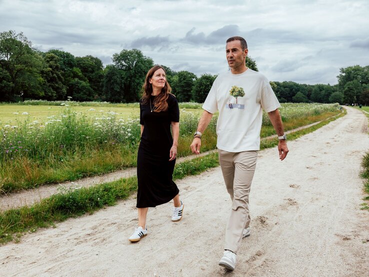 Eine Frau in schwarzem Kleid und ein Mann in hellem Shirt spazieren bei bewölktem Himmel auf einem Sandweg durch einen grünen Park. | © Maximilian Ralf Schmidt