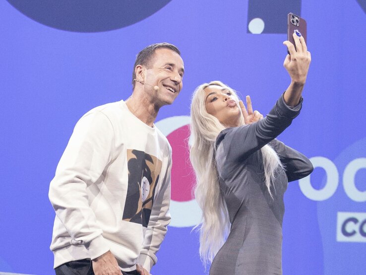 Ein Mann in weißem Pullover und eine blonde Frau in grauem Kleid machen lachend ein Selfie auf einer Bühne. | © dpa