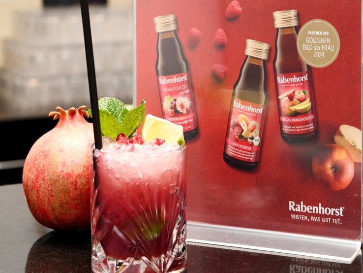 Ein fruchtiger Cocktail mit Granatapfelkernen, Minze und Limette steht vor einem roten Werbeaufsteller von Rabenhorst auf einer dunklen Theke.