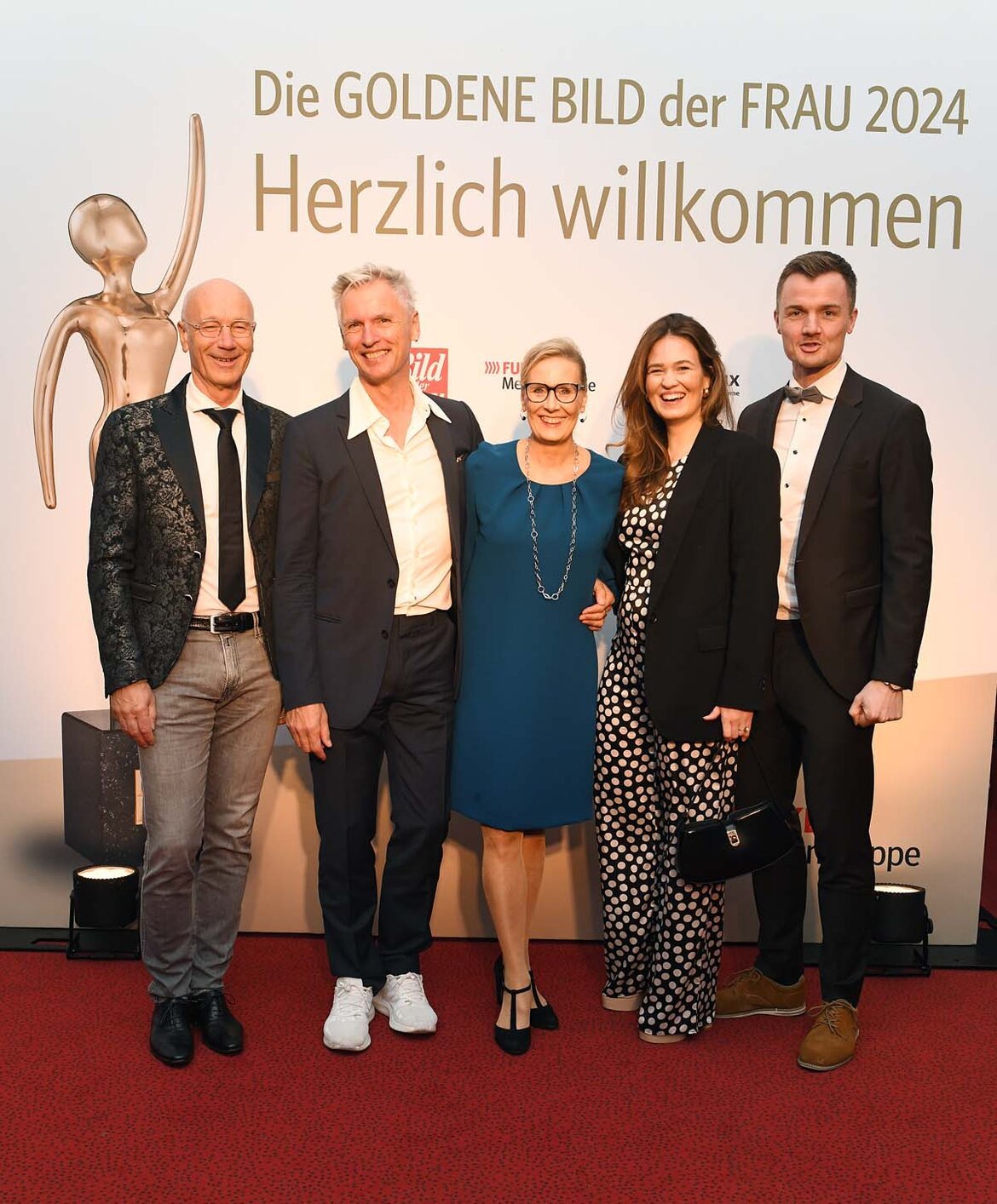 Fünf elegant gekleidete Personen stehen lächelnd auf einem roten Teppich vor einer Wand mit dem Schriftzug "Goldene Bild der Frau 2024" | © BILD der FRAU 