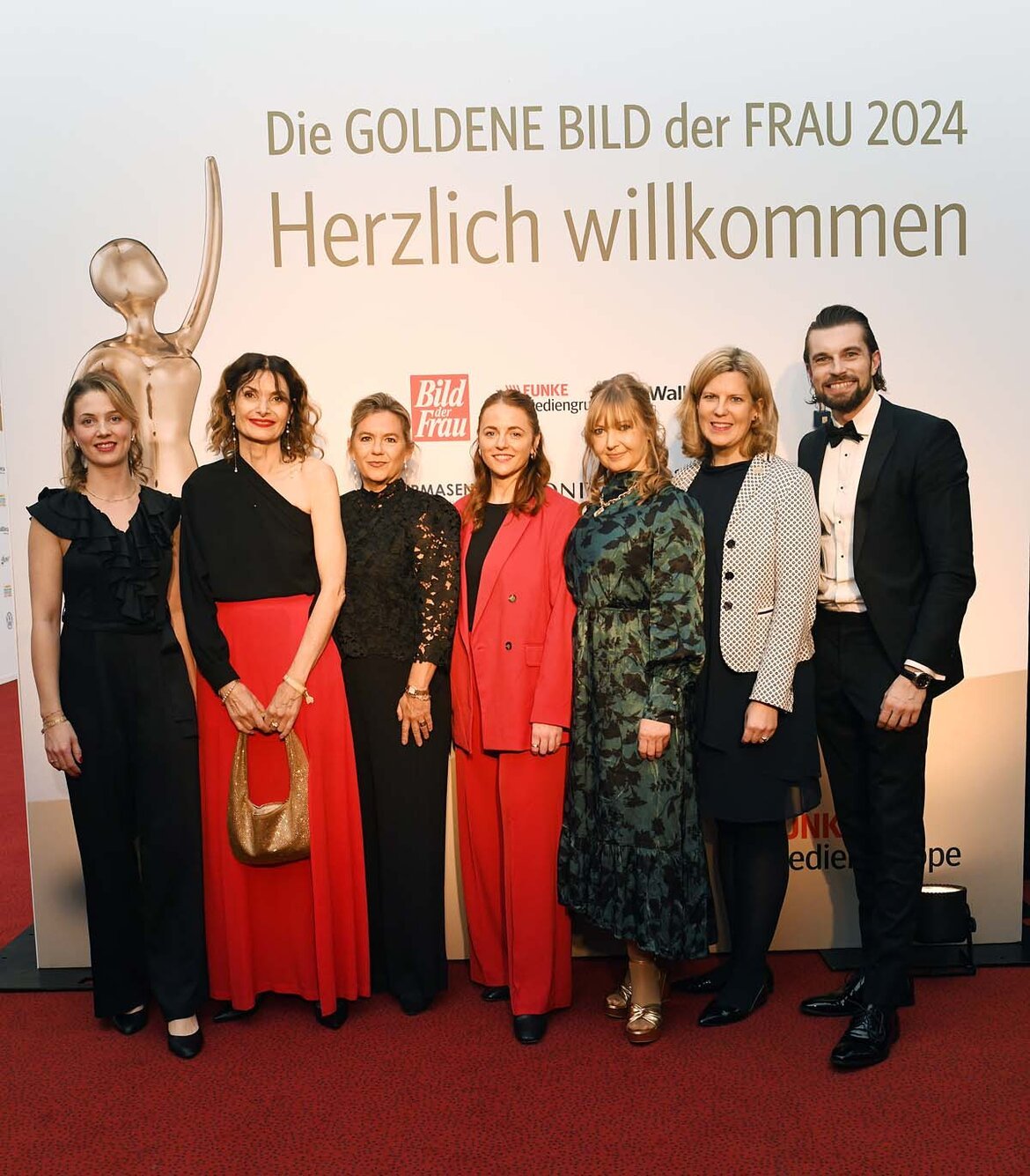 Sieben festlich gekleidete Personen lächeln auf einem roten Teppich vor einer Wand mit Schriftzug "Goldene Bild der Frau 2024 | © BILD der FRAU 