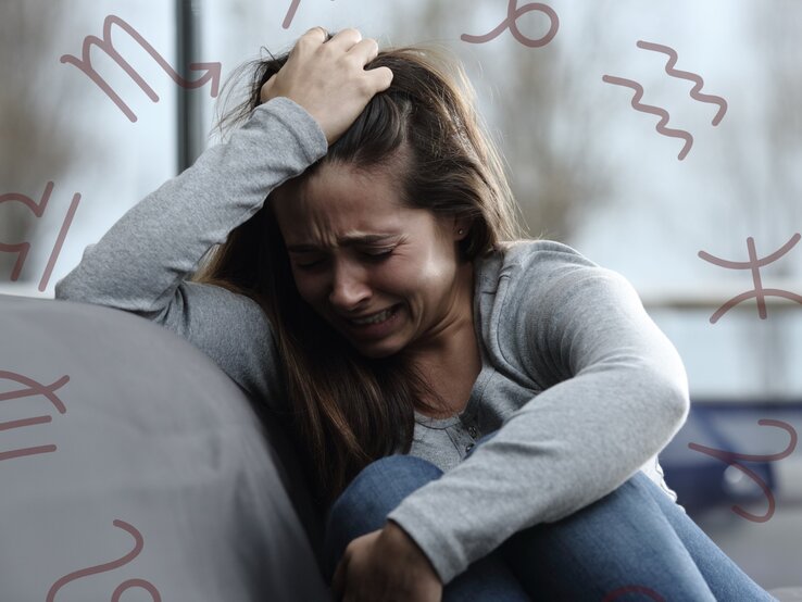 Eine junge Frau sitzt weinend und verzweifelt auf einem Sofa. Sie greift sich mit einer Hand in die Haare und wirkt sehr emotional und gestresst. Um sie herum sind verschiedene astrologische Symbole in einem dezenten Braunton zu sehen. Der Hintergrund ist unscharf und zeigt Bäume und ein Fenster, wodurch eine bedrückende Stimmung erzeugt wird.