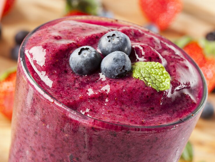 Ein dichter, lila-roter Smoothie in einem Glas, garniert mit frischen Blaubeeren und einem Minzblatt. Im Hintergrund sind verschwommene Erdbeeren und Blaubeeren zu sehen.