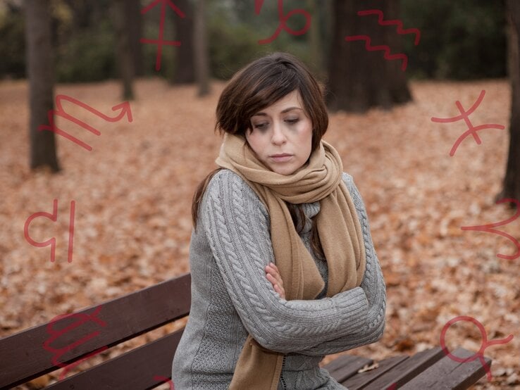 Eine nachdenkliche Frau sitzt mit verschränkten Armen auf einer Parkbank an einem Herbsttag. Sie trägt einen grauen Strickpullover und einen beigen Schal. Um sie herum liegen gefallene Blätter. Im Hintergrund sind Symbole von Sternzeichen zu sehen, die in Rot über das Bild gelegt sind. Die Frau wirkt bedrückt oder nachdenklich, möglicherweise in Verbindung mit einer Herbstdepression.