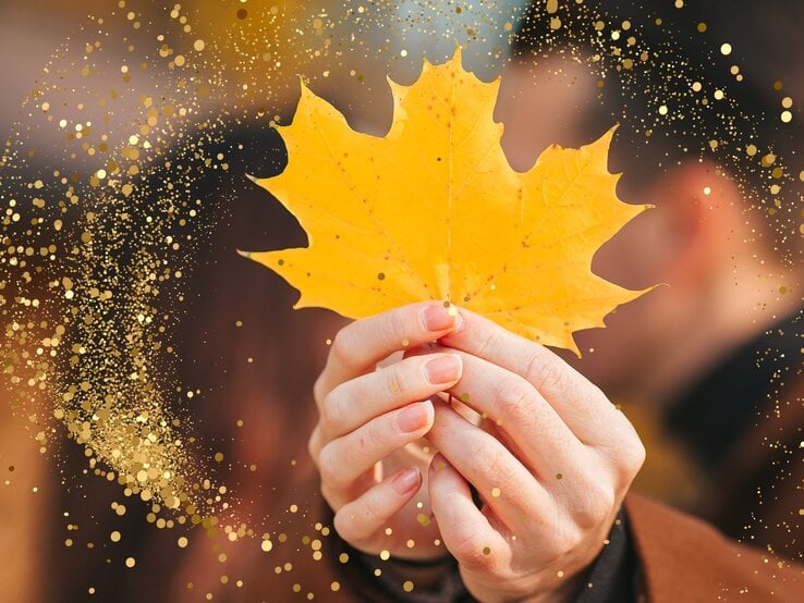 Ein herbstliches Bild mit einem goldgelben Ahornblatt, das von zwei Händen gehalten wird. Im Hintergrund ist unscharf eine Person zu erkennen. Goldene Glitzer- und Lichtpunkte sind über das Bild verteilt, was eine warme, magische Atmosphäre erzeugt.