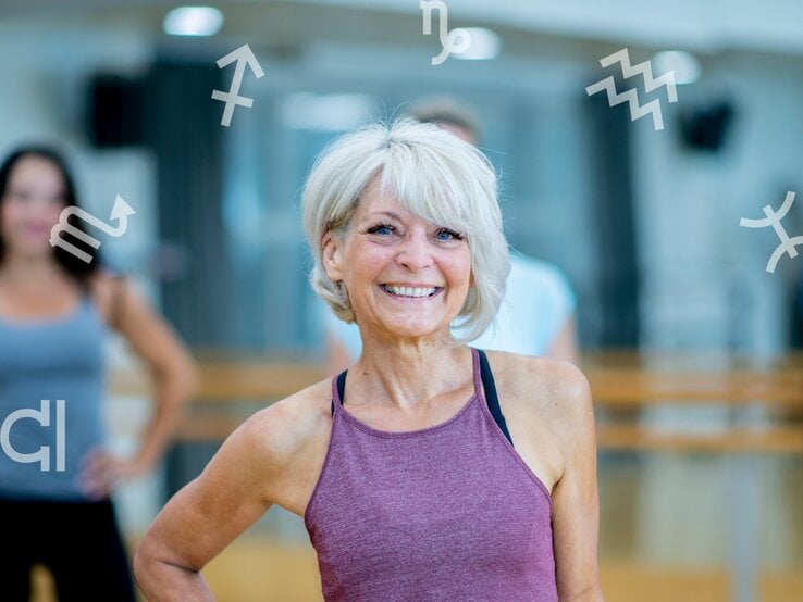 Eine ältere, sportliche Frau mit grauem Haar und lila Sporttop lächelt selbstbewusst in die Kamera. Sie befindet sich in einem hellen Fitnessstudio. Im Hintergrund sind unscharf weitere Personen zu sehen. Um sie herum schweben verschiedene astrologische Sternzeichen-Symbole, was auf einen Zusammenhang zwischen Fitness und Astrologie hinweist.