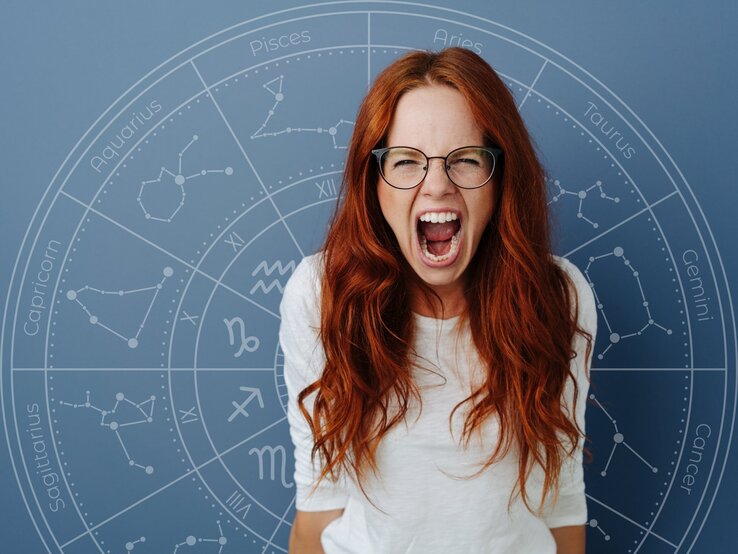 Eine rothaarige Frau mit Brille schreit wütend oder frustriert in die Kamera. Im Hintergrund ist ein astrologischer Kreis mit den Sternzeichen-Symbolen auf einem blauen Hintergrund zu sehen.