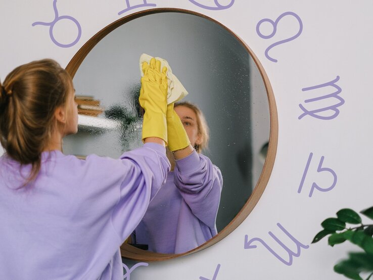 Eine junge Frau mit braunen Haaren, die zu einem lockeren Zopf gebunden sind, trägt einen lila Pullover und gelbe Gummihandschuhe, während sie einen runden Spiegel mit einem Tuch reinigt. Ihr Spiegelbild ist gut sichtbar. Der Hintergrund ist eine helle Wand, auf der astrologische Sternzeichen in lila gezeichnet sind. Die Szene vermittelt ein Gefühl von Ordnung und Reinigung, sowohl im physischen als auch im symbolischen Sinne.