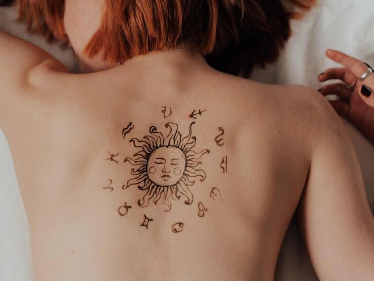 Eine Person mit roten Haaren liegt auf dem Bauch, während ihr Rücken ein temporäres oder permanentes Tattoo mit einer Sonne und astrologischen Symbolen ziert. Die Sonne im Zentrum des Designs hat ein Gesicht und wird von den zwölf Tierkreiszeichen umgeben. Die Person trägt Schmuck an der Hand und befindet sich in einer entspannten Haltung.