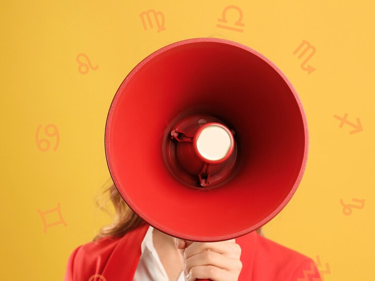 Ein Person hält ein großes rotes Megafon vor ihrem Gesicht. Im Hintergrund sind verschiedene Sternzeichen-Symbole auf einem gelben Hintergrund zu sehen. Die Person trägt eine rote Jacke und ein weißes Hemd.