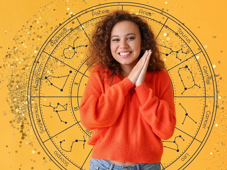 Eine junge Frau mit lockigem braunen Haar und einem orangefarbenen Pullover lächelt fröhlich und hält ihre Hände vor dem Gesicht zusammen. Hinter ihr ist ein großer astrologischer Tierkreis auf einem gelben Hintergrund zu sehen, der die Sternzeichen und ihre Konstellationen darstellt. Goldene Glitzerpunkte verstärken die visuelle Wirkung des Bildes.