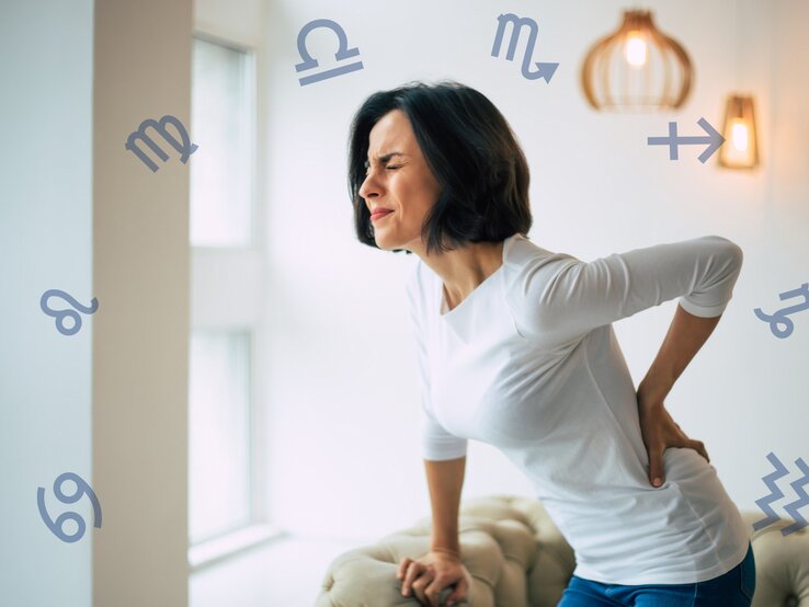 Eine Frau mit kurzen dunklen Haaren hält sich mit schmerzverzerrtem Gesicht den unteren Rücken, während sie sich an einer Couch abstützt. Um sie herum schweben verschiedene astrologische Symbole der Sternzeichen, was auf einen Zusammenhang zwischen Rückenschmerzen und den Sternzeichen hinweist.